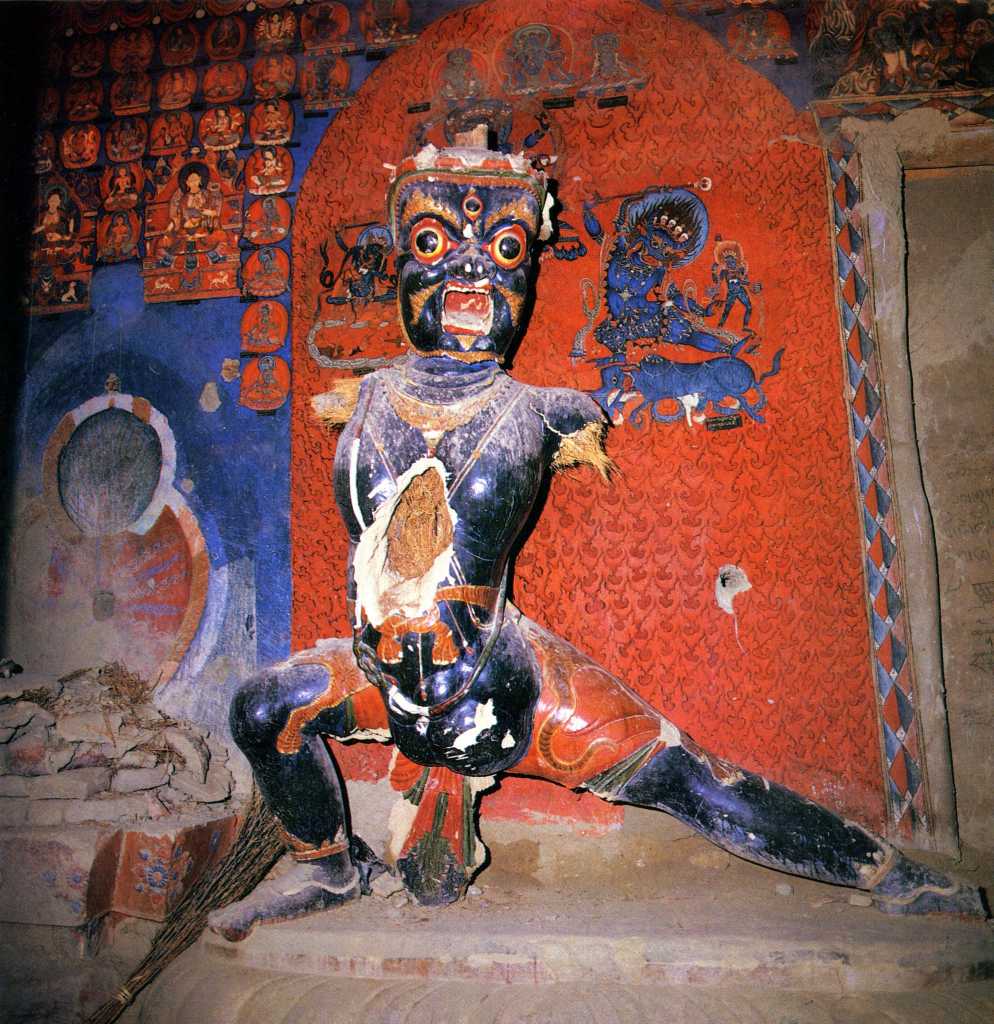 Tibet Guge 06 Tsaparang White Temple 05 23 Vajrapani Immediately to the right of the door is a 5m-high guardian figure, blue Vajrapani (Tib. Chana Dorje). The significantly elongated torsos, a typical Guge trademark, have been broken open, exposing the straw. This has since been closed with what looks like white plaster. To the left of Vajrapani is an empty pedestal that used to contain a statue of Tara. Photo - Weyer/Aschoff: Tsaparang, Tibets Grosses Geheimnis.